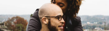 A woman with eyes closed embracing a man wearing glasses from behind